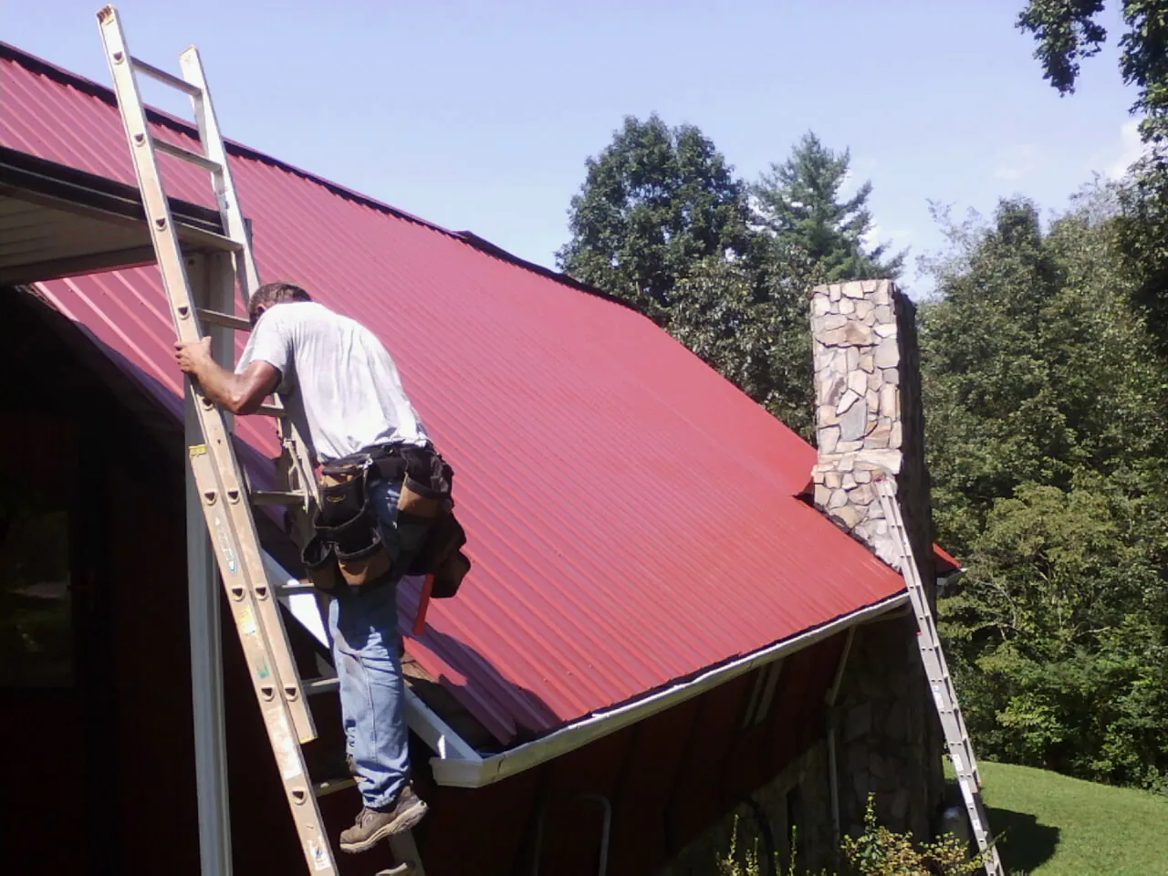 Roofer on a ladder beside a red metal roof and stone chimney — Soffit & Fascia Repair by Roofing Brothers in Foristell